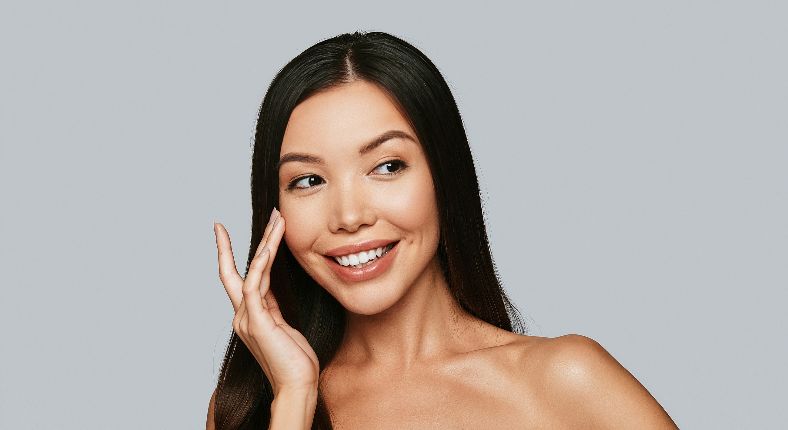 Smiling Melbourne Botox model touching her cheek, gray background.